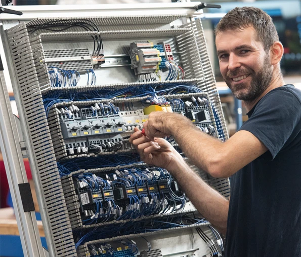 harich Elektriker verkabelt in offenem Schaltschrank, dreht sich lächelnd zur Kamera und bietet Einblick in die Elektronik