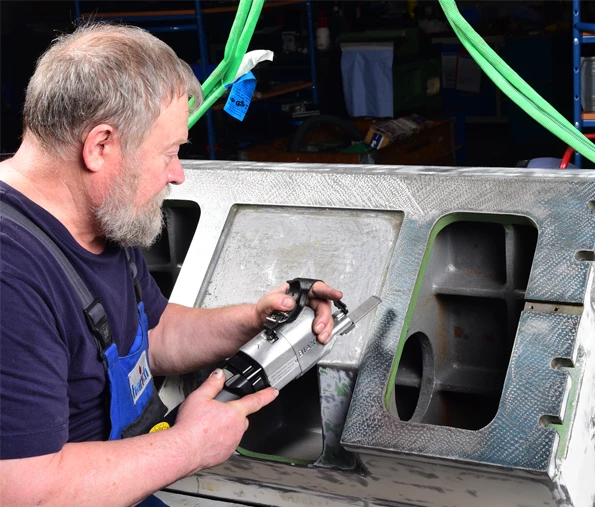 Mann sitzt seitlich und schabt manuell ein großes Maschinenteil mit einem Elektroschaber, trägt harich Arbeitskleidung
